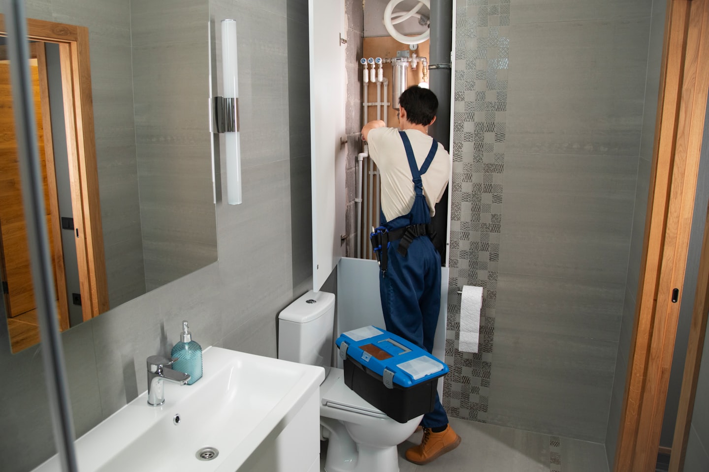 Tecnico ripara idraulica dietro il water in un bagno con piastrelle grigie.